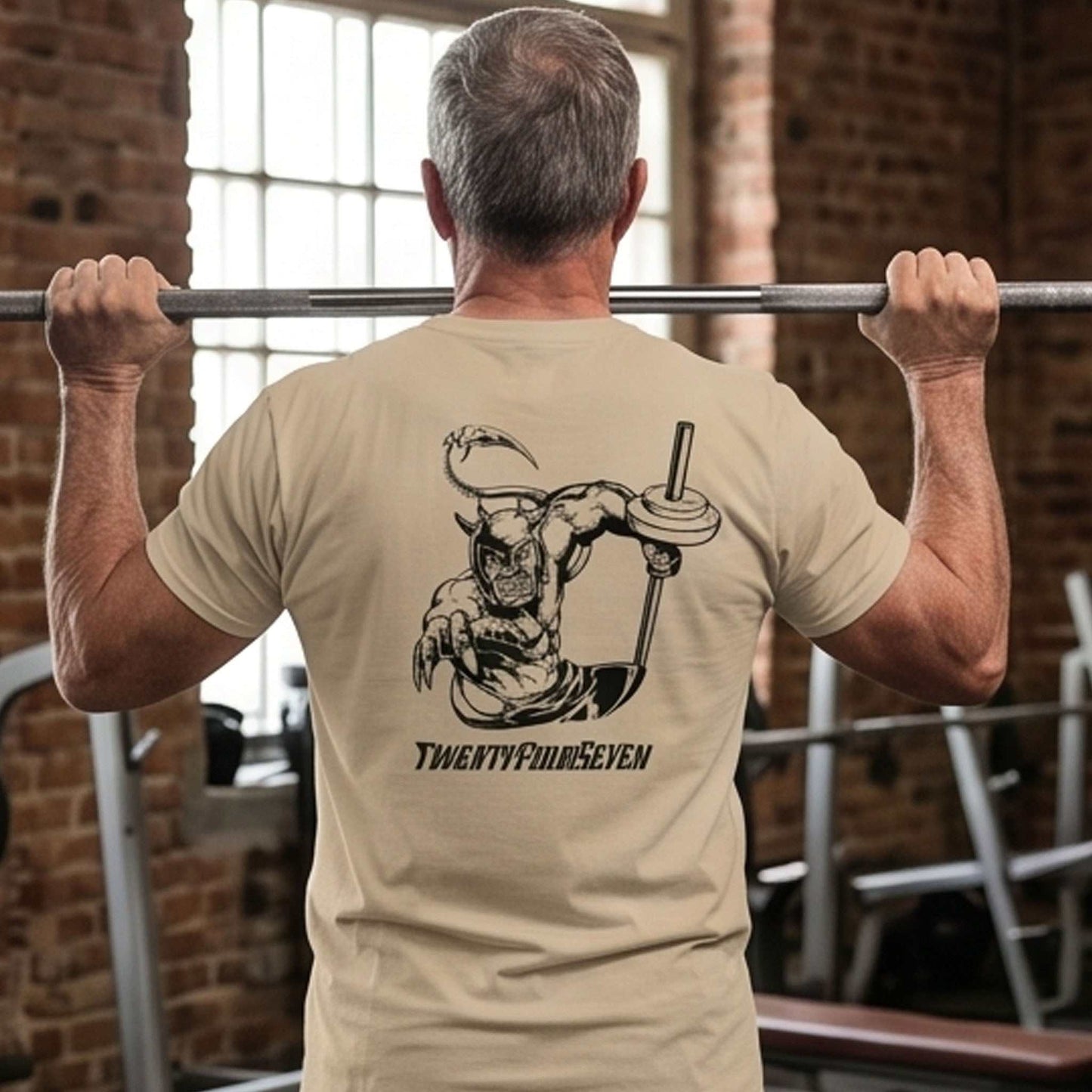Muscular male model seen from the back wearing the sand beige unisex bodybuilding graphic tee, preparing to shoulder press with a barbell in a rustic brick-wall gym, highlighting the Dablo back graphic.