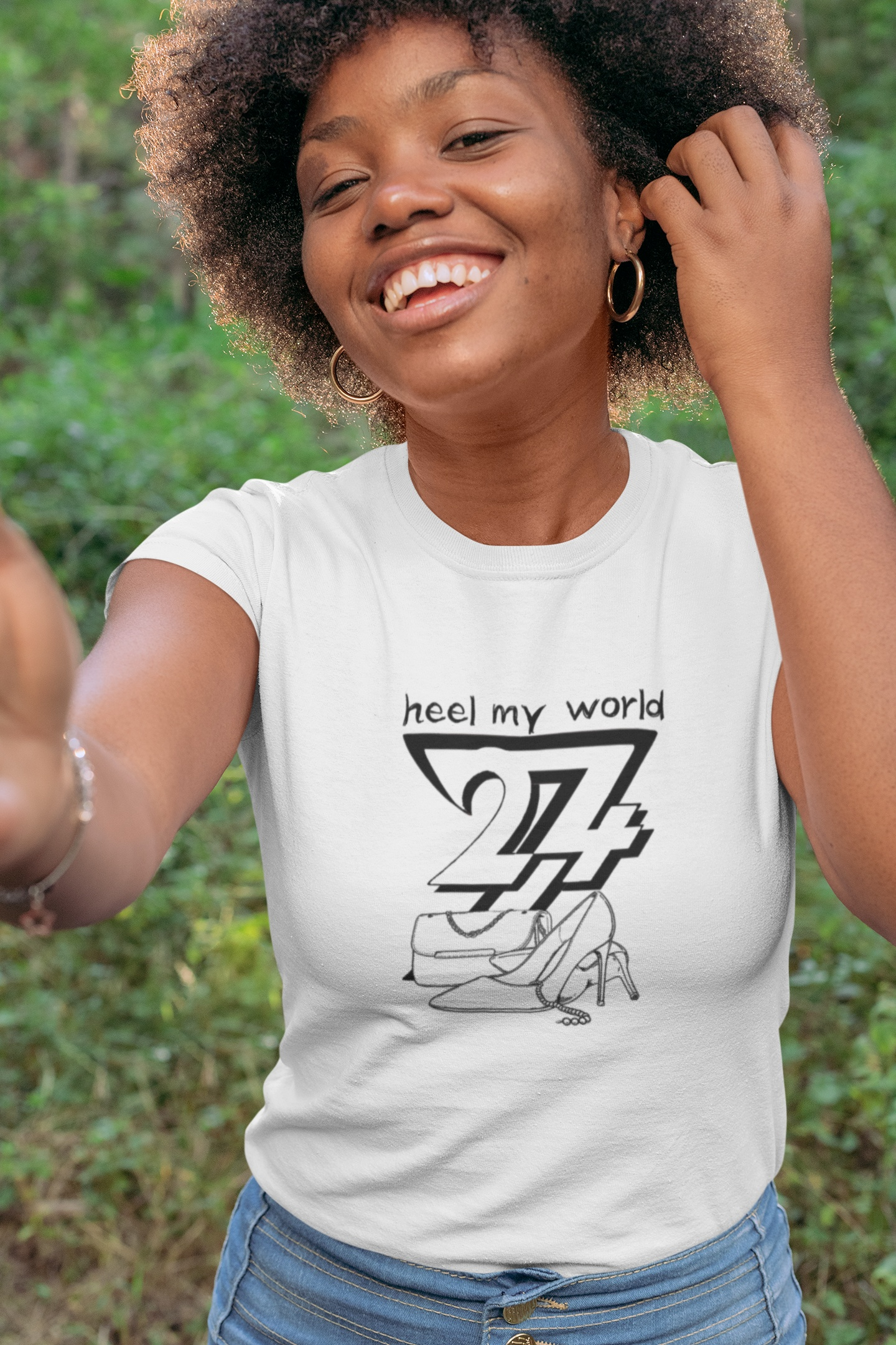 A smiling female model standing outdoors in a natural woodland setting, wearing the white minimalist Heel My World graphic tee.