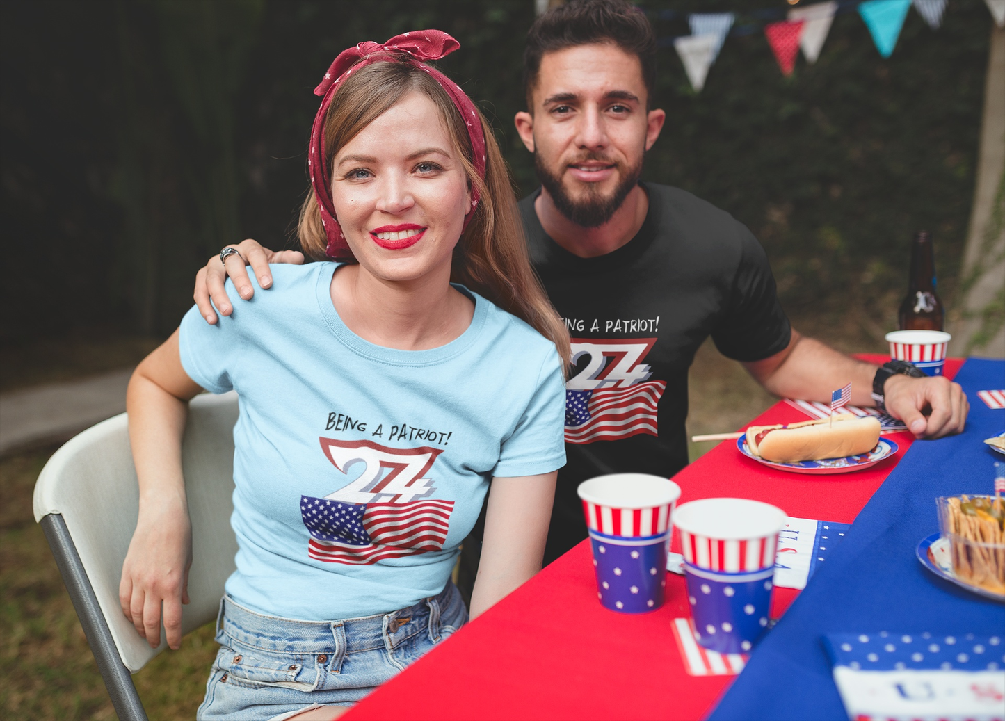 Female model in the light blue patriotic rally gear tee and a male model in the black variant, smiling happily together at a festive outdoor 4th of July BBQ.