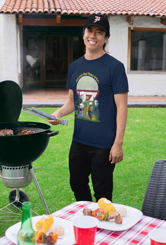 Man wearing a navy blue funny BBQ t-shirt with a father and son cactus camping graphic, paired with a black baseball cap featuring the 24/7 logo.
