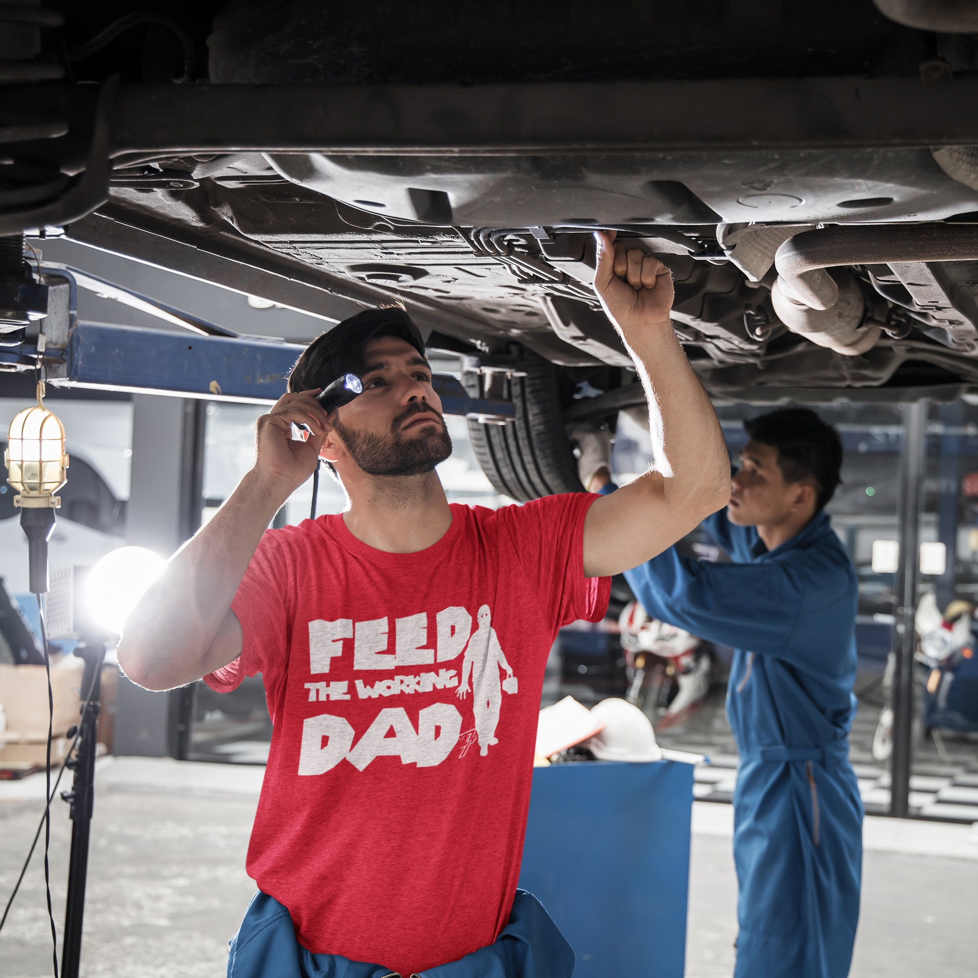 Male model wearing the red mechanic Father's Day shirt featuring the crisp white zombie tradesman graphic, inspecting a car in an auto repair shop.
