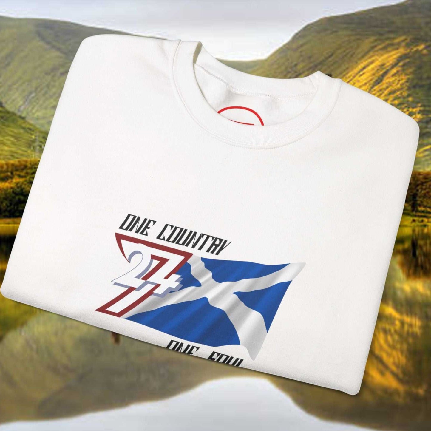 White Scotland Saltire crewneck sweatshirt with hand-drawn St. Andrew’s Cross design and "One Country One Soul" text, folded on a scenic Scottish landscape background