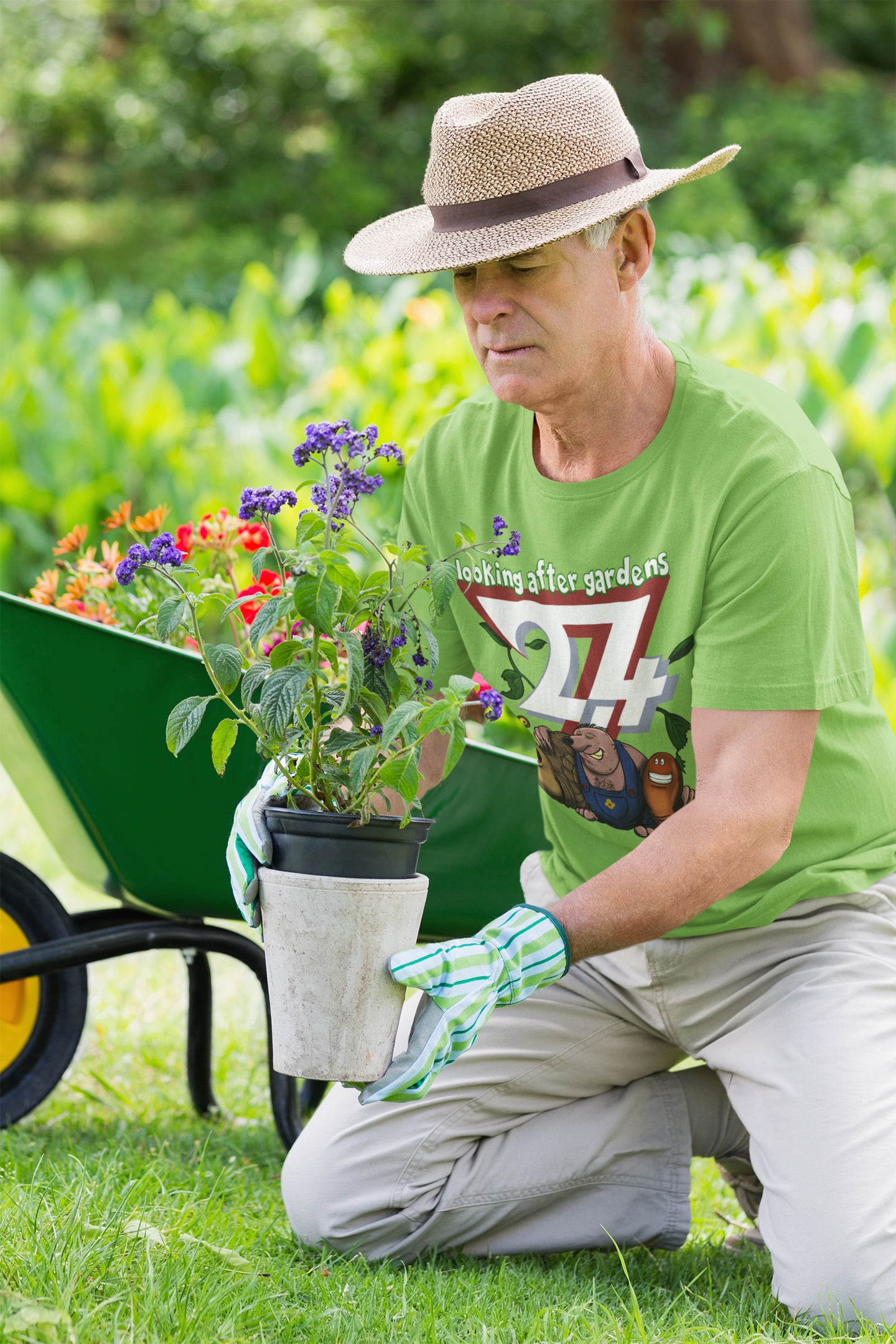 Senior male model wearing the bright green plant lover t-shirt and a sun hat, carefully planting purple flowers near a green wheelbarrow.