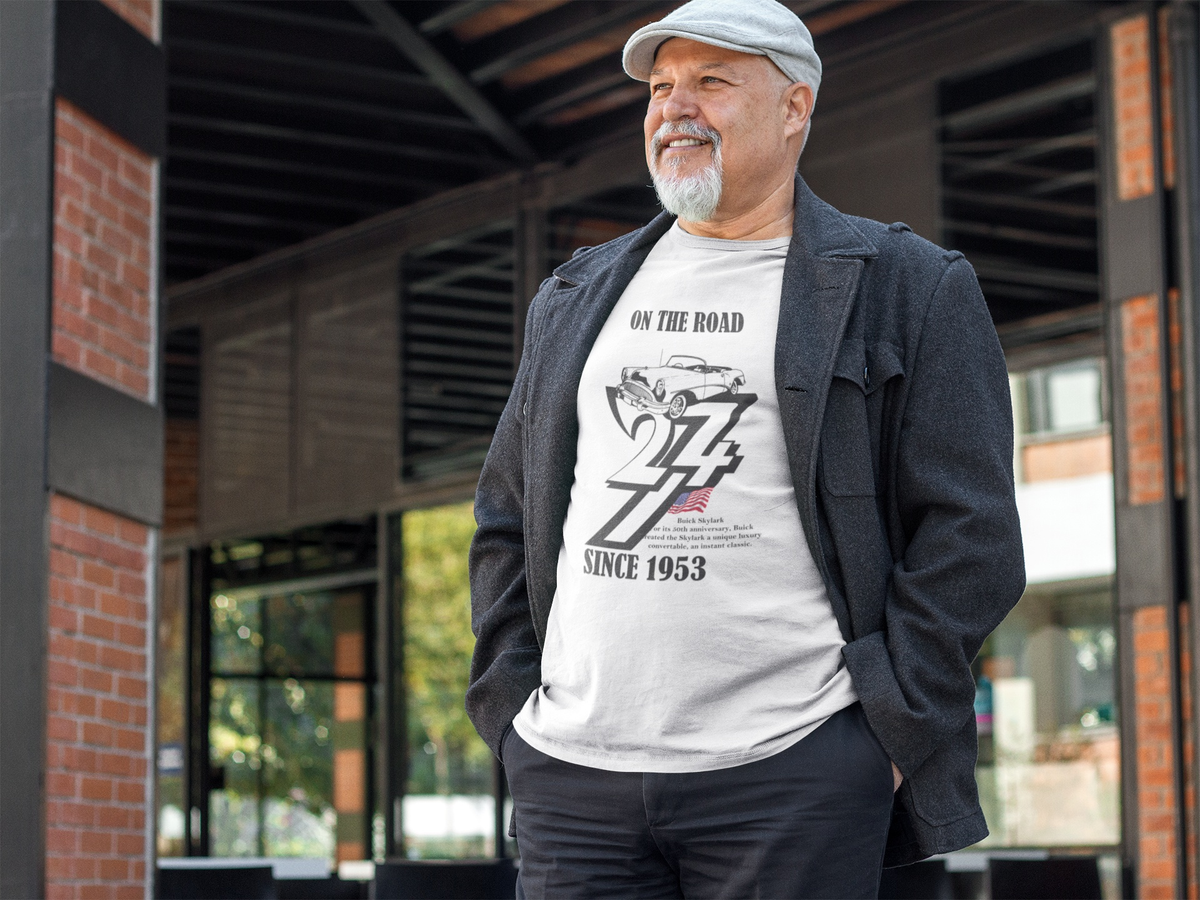 Smiling senior man with a goatee and newsboy cap standing outside a cafe wearing the white 1953 Buick Skylark graphic tee under a charcoal jacket.
