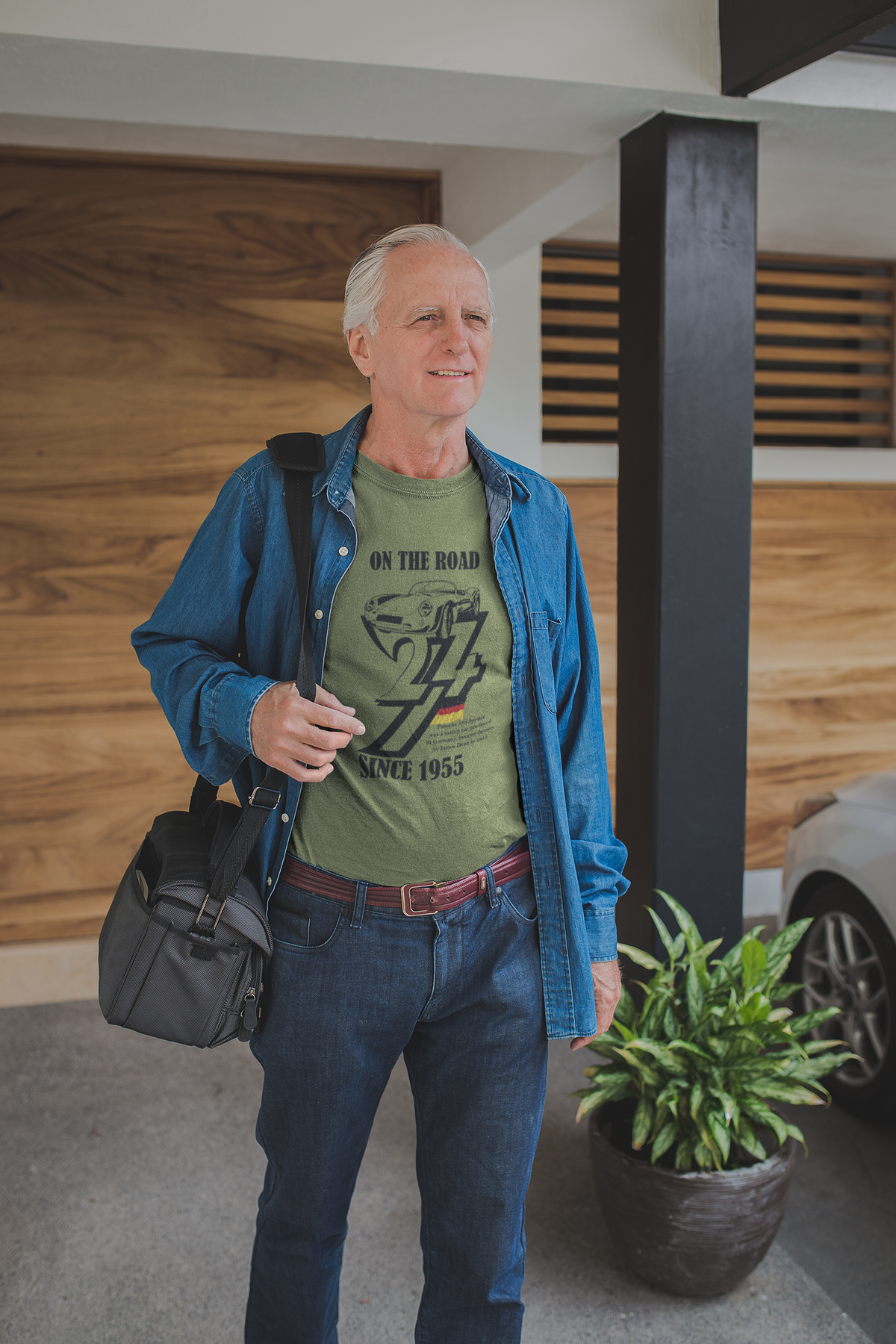 Handsome senior man with gray hair wearing the army green 1955 vintage Porsche tee under a denim overshirt, holding a duffel bag while walking out of a modern home.