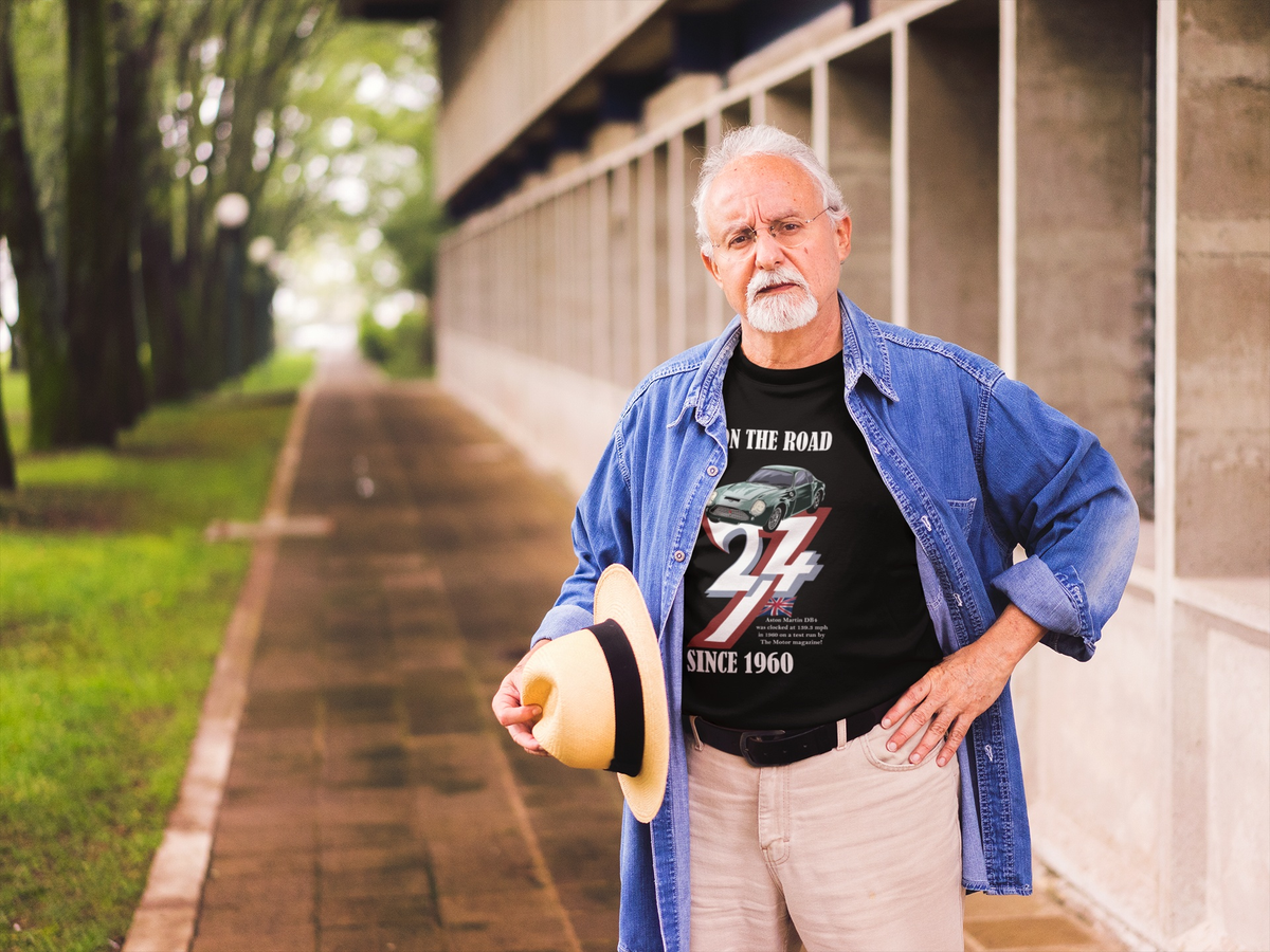 Distinguished senior man with a white goatee wearing an open denim shirt over the black classic car graphic tee, holding a straw hat outdoors near a garden pathway.
