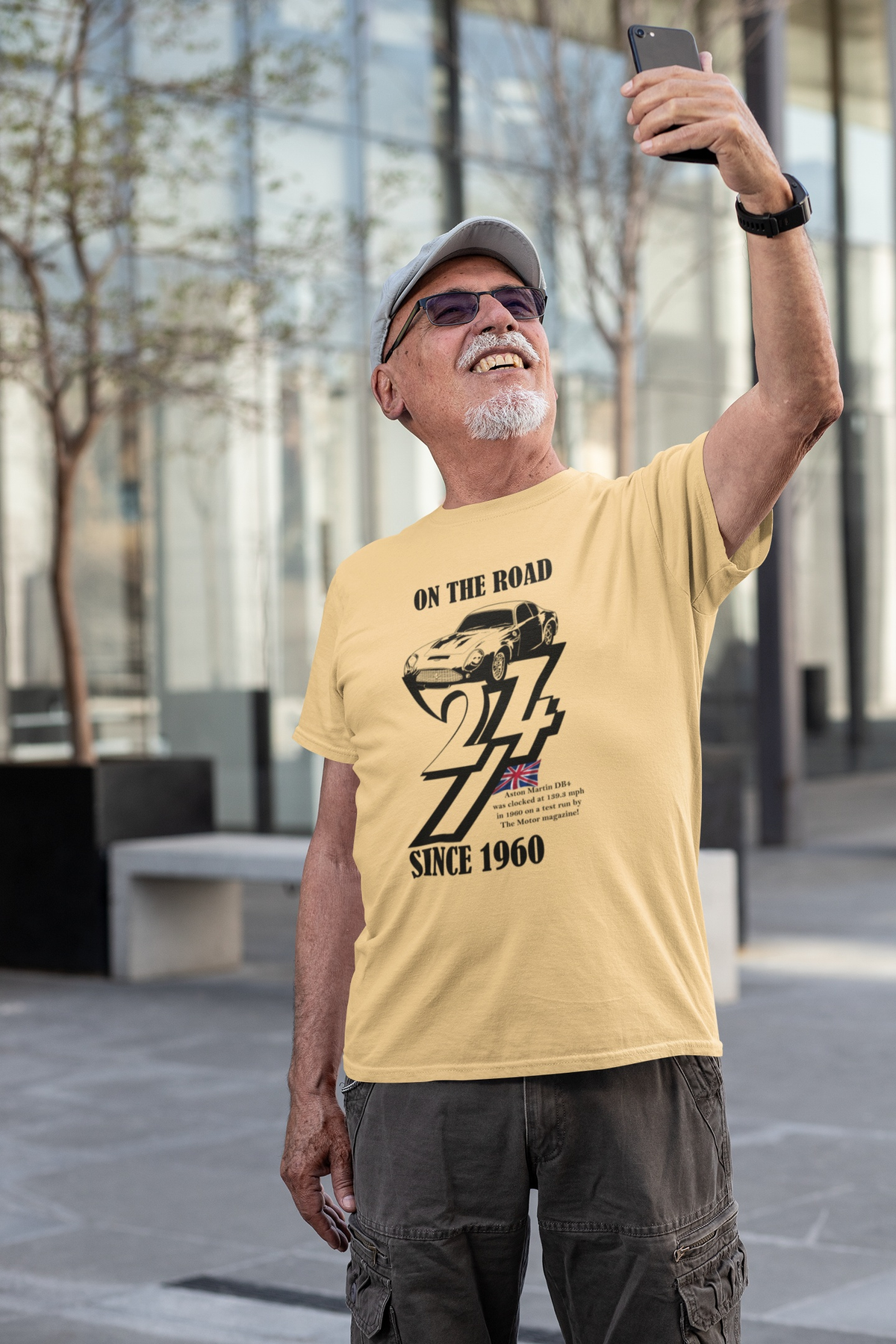 Smiling senior man with a goatee and baseball cap taking a selfie outdoors in a modern city plaza, wearing the yellow haze 1960 Aston Martin vintage tee.