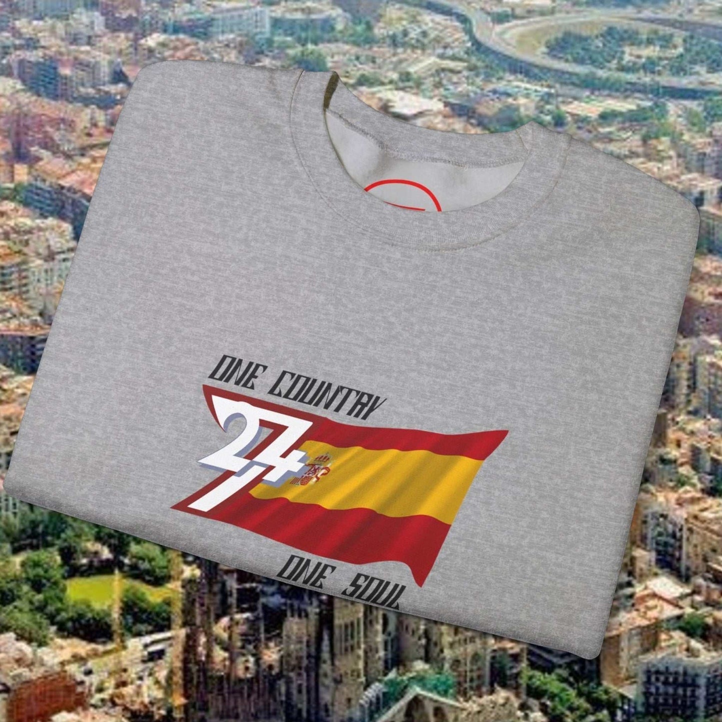Grey Spain flag sweatshirt with hand-drawn graphic and text "One Country One Soul" folded on cityscape background