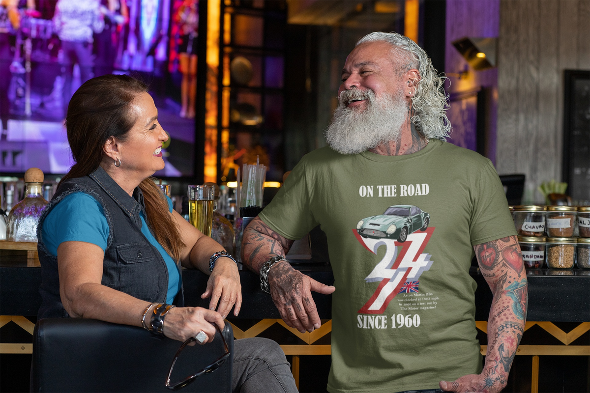 Smiling senior man with a thick white beard and arm tattoos sitting at a bar chatting with a woman, wearing the army green 1960 Aston Martin graphic tee.