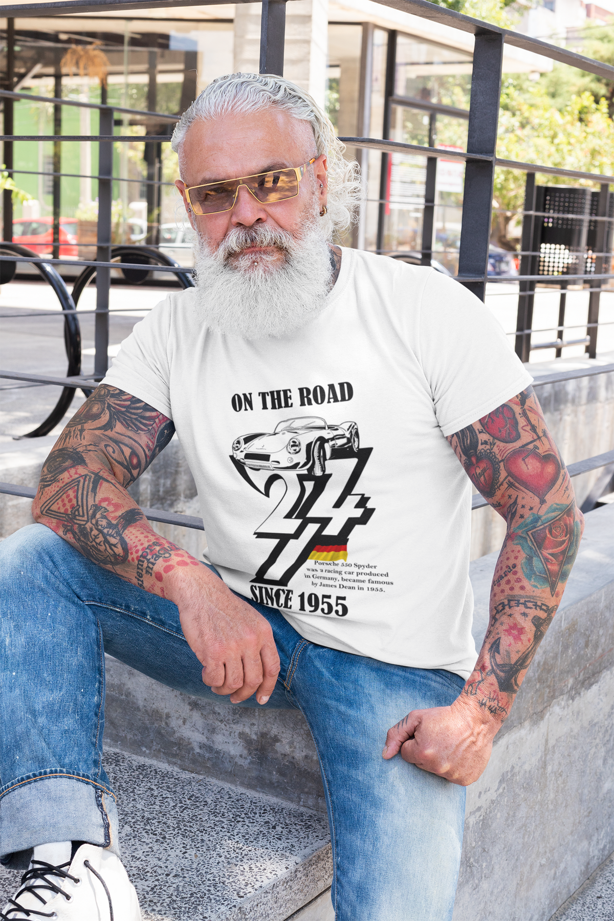 Stylish senior man with a full white beard, sunglasses, and tattooed arms sitting outdoors on concrete steps, modeling the white born in 1955 classic car birthday tee.