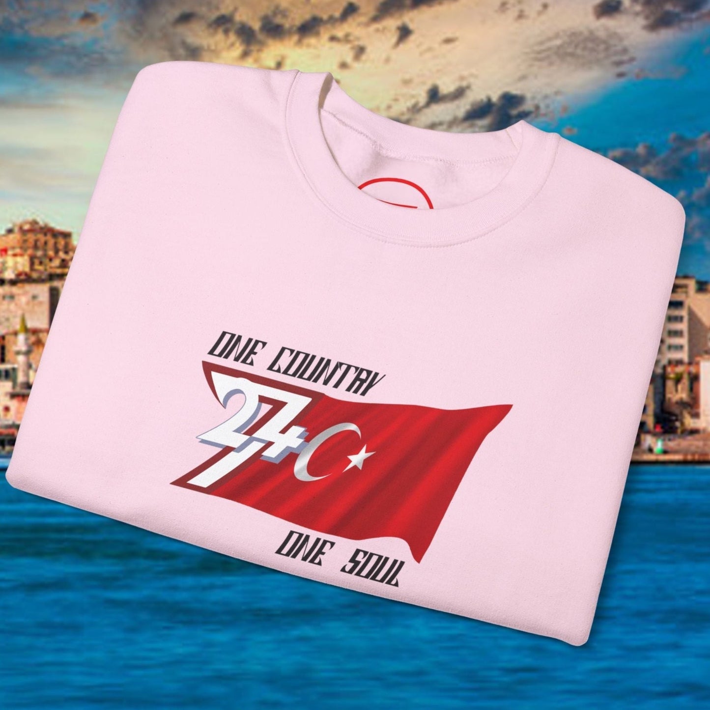 Folded pink Turkish Flag Sweatshirt with hand-drawn design featuring crescent and star and text "One Country One Soul" on waterfront background