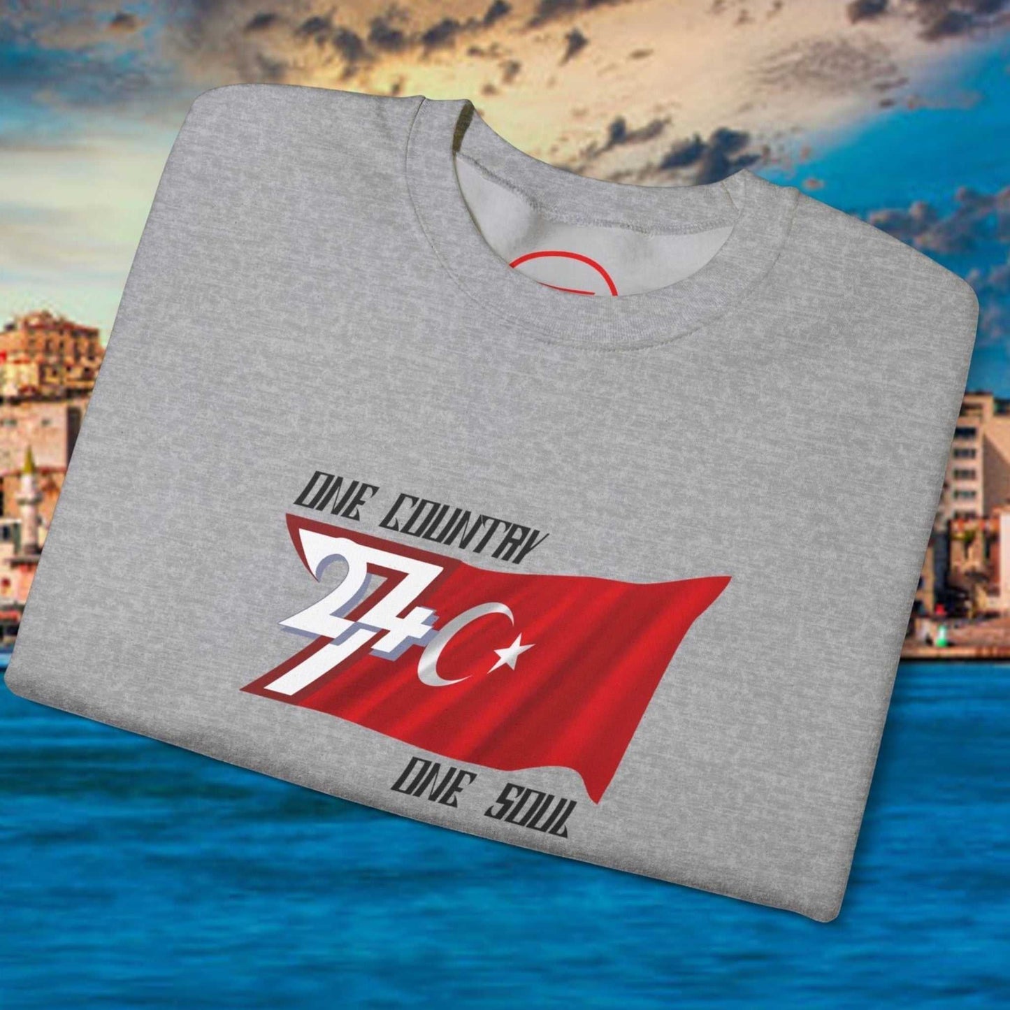 Folded gray sweatshirt with hand-drawn Turkish flag design and text one country one soul against coastal city backdrop