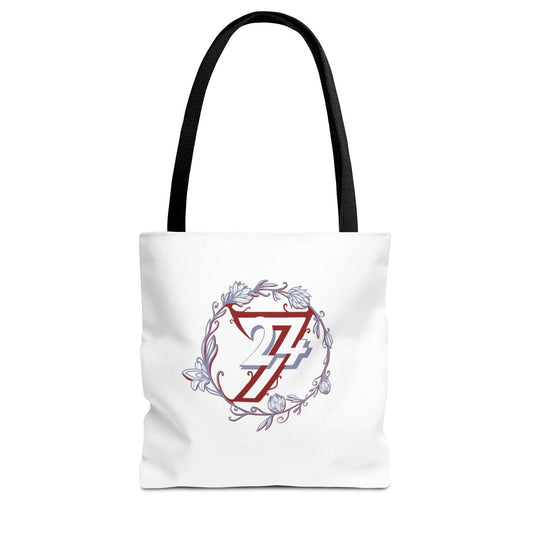 A white canvas floral tote bag with black shoulder straps, featuring a central graphic of a red stylized 24/7 inside a circular floral wreath with blue and gray accents.