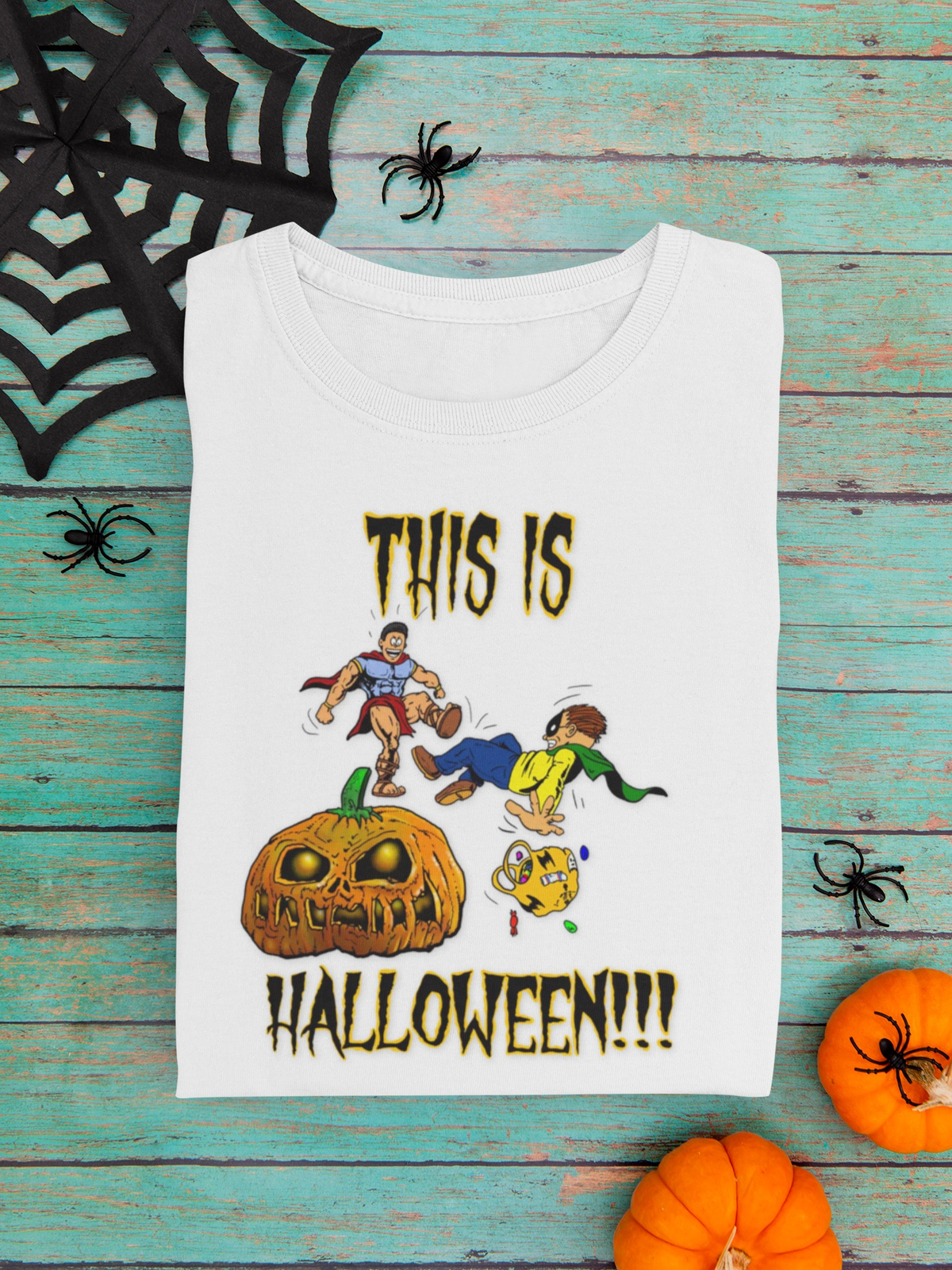 A white short-sleeve T-shirt laid flat on a distressed turquoise wood surface, printed with the phrase 'THIS IS HALLOWEEN!!!' in black outlined letters and a Halloween-themed cartoon scene featuring a pumpkin, a ghost, and two children. Decorative black spiderwebs and spiders surround the shirt, with small orange pumpkins at the bottom corners.