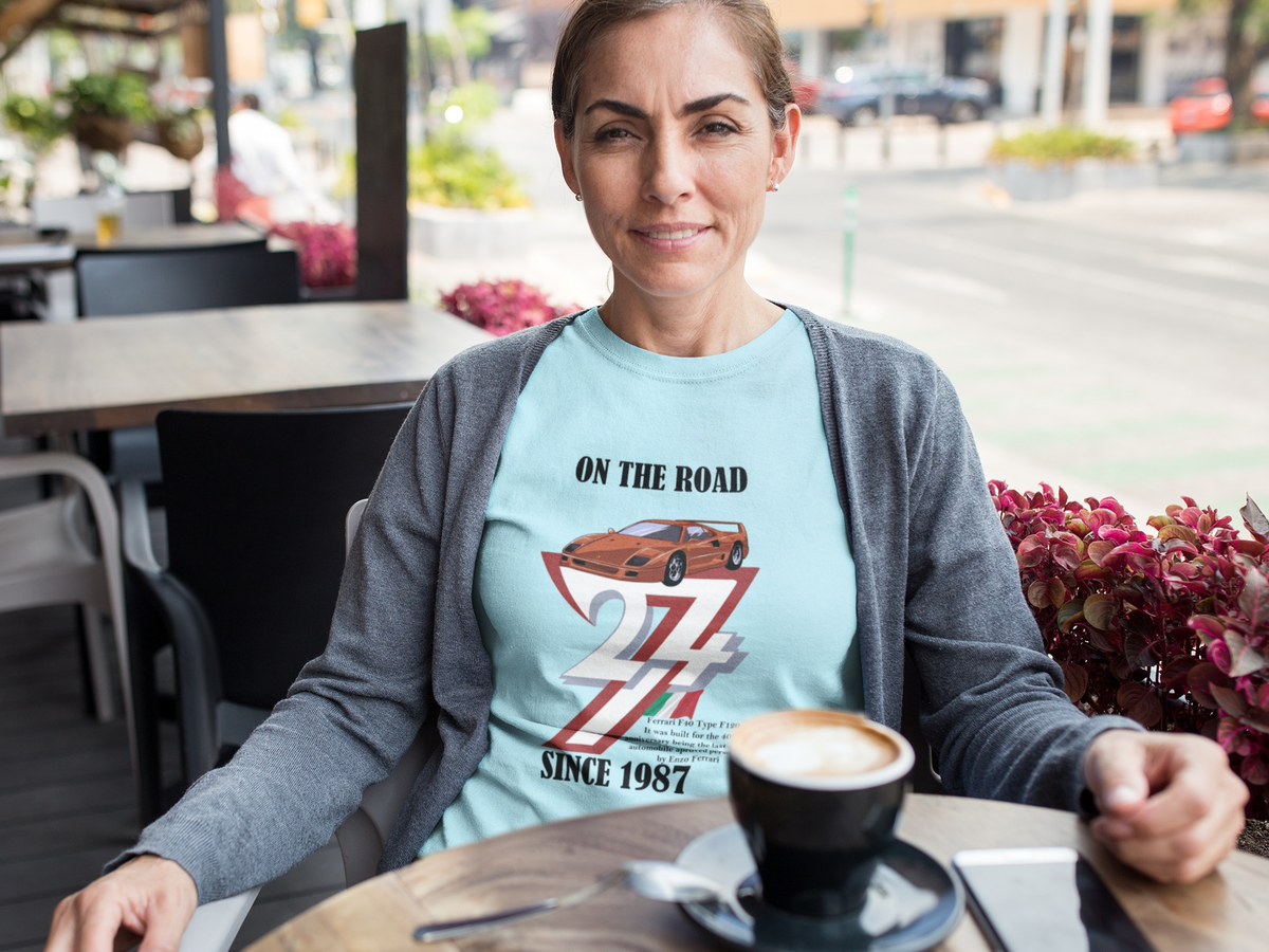 Smiling middle-aged woman wearing a gray cardigan over the light blue born in 1987 t-shirt, sitting at an outdoor cafe table with a cappuccino.
