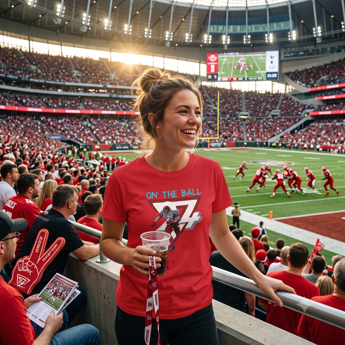 Joyful young woman standing in a crowded football stadium holding a beverage, wearing the bold red hand-drawn game day shirt as players compete on the field below.