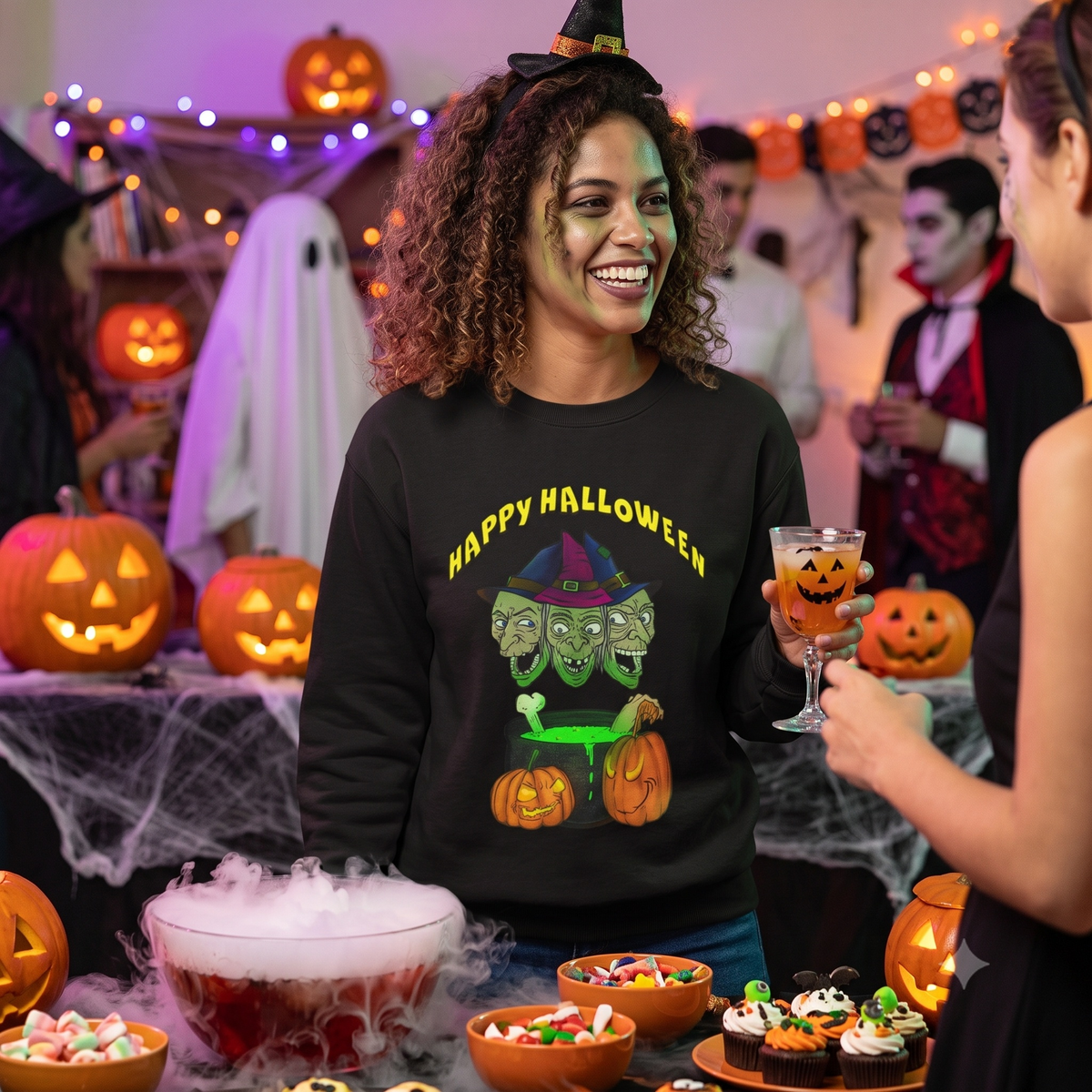 Smiling young woman enjoying a lively indoor Halloween party holding a festive drink, wearing the black spooky Halloween sweatshirt featuring the front witches graphic.