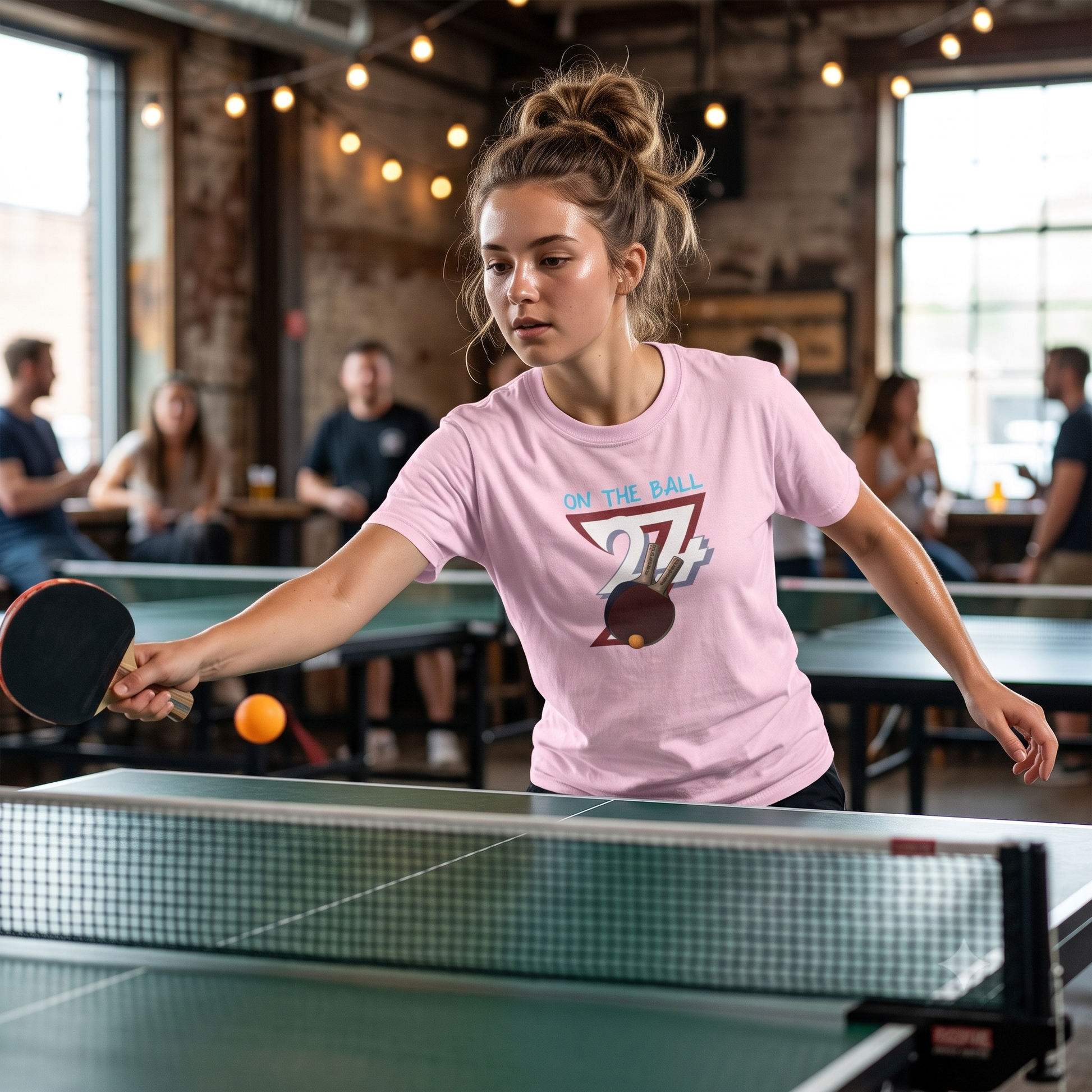 Concentrating young woman serving a ping pong ball during a friendly indoor club game, wearing the light pink table tennis graphic tee.