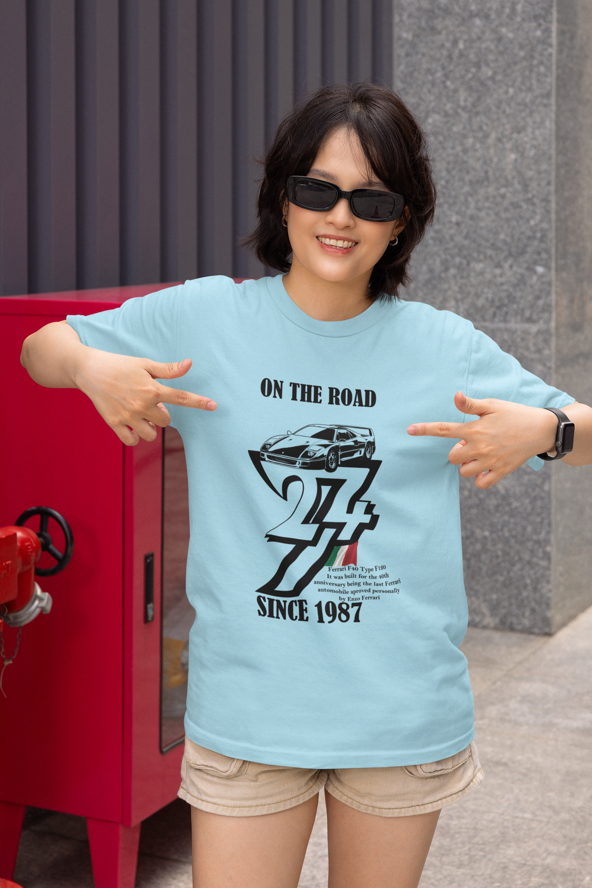Smiling woman wearing sunglasses outside near a red utility box, pointing enthusiastically at the black vintage Ferrari F40 graphic on her light blue t-shirt.