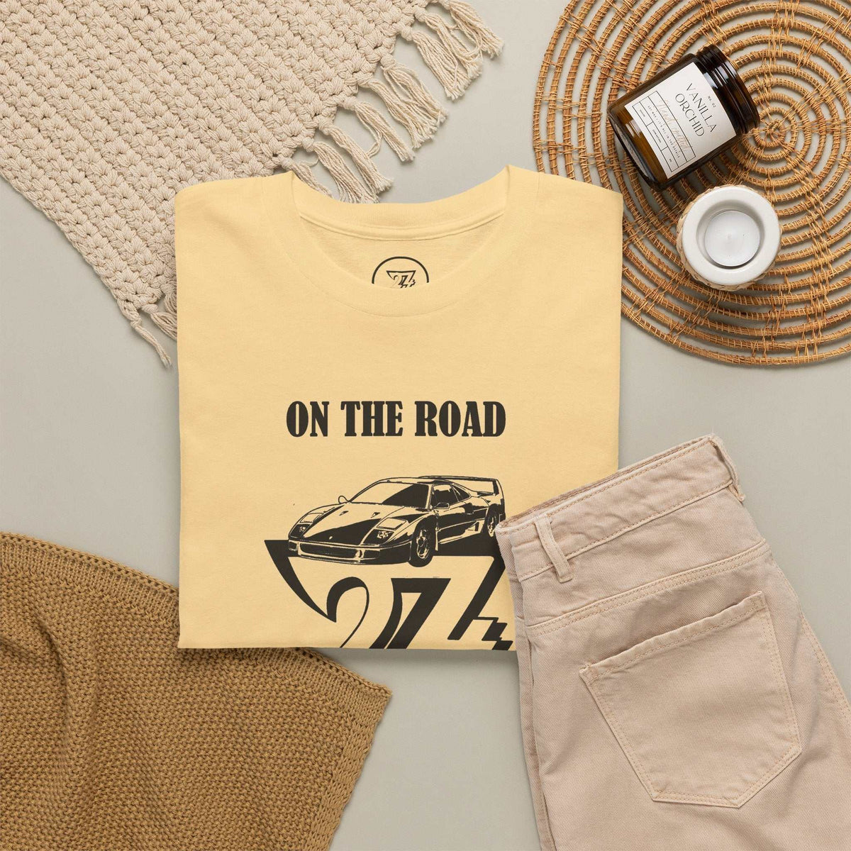 A folded pale yellow T-shirt with a black 'ON THE ROAD' text and sports car graphic, styled in a flat lay with beige pants, a woven blanket, a candle jar, and a small cup on a neutral background.